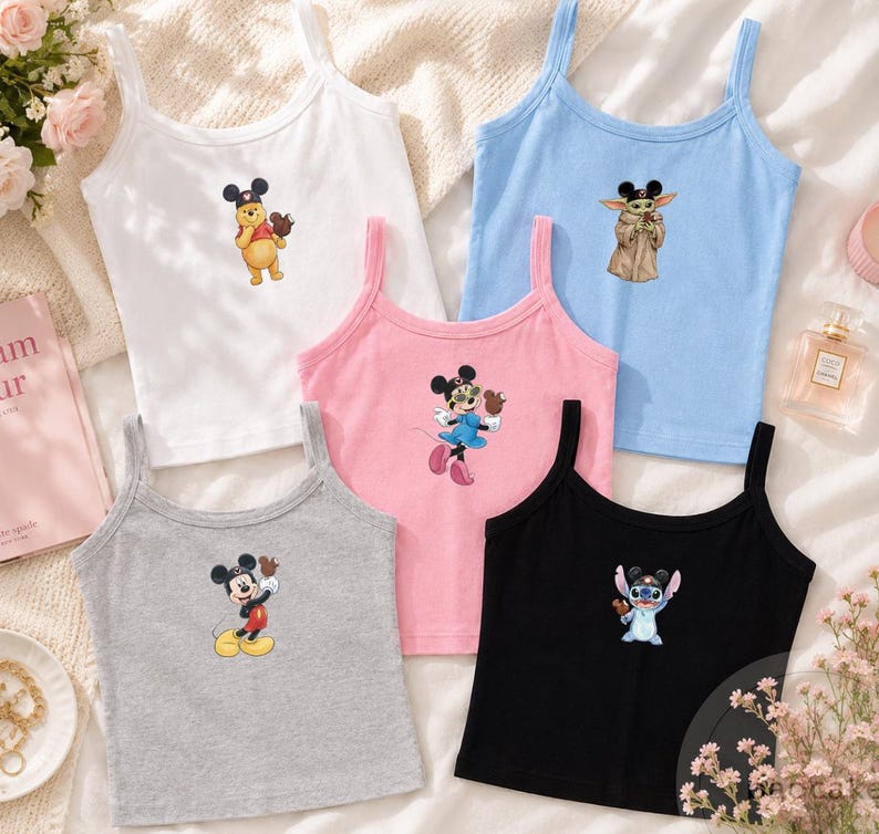 May include: A collection of five sleeveless tank tops in white, light blue, pink, gray, and black. Each top features a small graphic print of a different Disney character wearing Mickey Mouse ears, including Winnie the Pooh, Grogu, Minnie Mouse, Mickey Mouse, and Stitch.