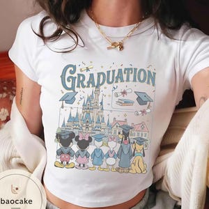 May include: White t-shirt featuring a colorful graphic with the word "Graduation" in blue, a castle, and cartoon characters wearing graduation caps. The characters include Mickey Mouse, Minnie Mouse, Donald Duck, Daisy Duck, and Pluto.
