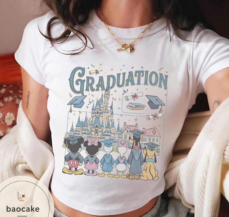 May include: White t-shirt featuring a colorful graphic with the word "Graduation" in blue, a castle, and cartoon characters wearing graduation caps. The characters include Mickey Mouse, Minnie Mouse, Donald Duck, Daisy Duck, and Pluto.