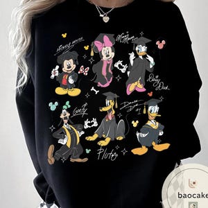 May include: Black sweatshirt featuring cartoon characters Mickey Mouse, Minnie Mouse, Daisy Duck, Donald Duck, Goofy, and Pluto in graduation attire. Each character is wearing a graduation cap and gown. The names of the characters are written above or below them.