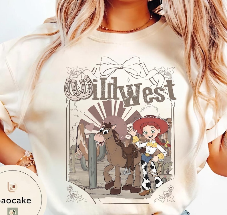 Pu&ograve; includere: T-shirt color crema con un design del West selvaggio. La grafica include una cowgirl dei cartoni animati, un cavallo e la scritta "Wild West" in stile vintage. Sono inclusi anche un ferro di cavallo e un cactus.