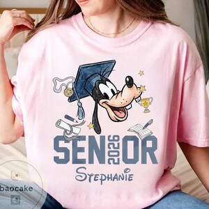 May include: Pink t-shirt featuring a cartoon character wearing a graduation cap and holding a trophy. The shirt has the text "SENIOR 2026" and a name below. The design includes stars, a diploma, and a book.