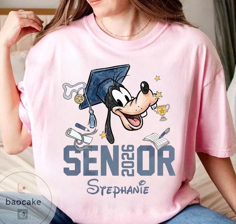 May include: Pink t-shirt featuring a cartoon character wearing a graduation cap and holding a trophy. The shirt has the text "SENIOR 2026" and a name below. The design includes stars, a diploma, and a book.