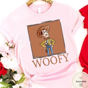 K&ouml;nnte beinhalten: Ein hellrosa T-Shirt mit kurzen &Auml;rmeln und einem quadratischen Aufdruck eines Cartoon-Cowboy-Charakters mit braunem Hut, gelbem Karohemd und blauer Hose. Unter dem Bild ist das Wort WOOFY in gro&szlig;en, fetten, braunen Gro&szlig;buchstaben aufgedruckt.