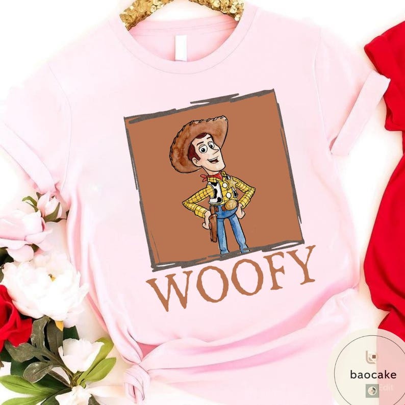 K&ouml;nnte beinhalten: Ein hellrosa T-Shirt mit kurzen &Auml;rmeln und einem quadratischen Aufdruck eines Cartoon-Cowboy-Charakters mit braunem Hut, gelbem Karohemd und blauer Hose. Unter dem Bild ist das Wort WOOFY in gro&szlig;en, fetten, braunen Gro&szlig;buchstaben aufgedruckt.