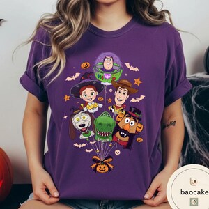 Pode incluir: Uma t-shirt roxa de manga curta com uma impress&atilde;o gr&aacute;fica de v&aacute;rias personagens animadas vestidas com fatos de Halloween, incluindo um ranger espacial, uma cowgirl, um cowboy, um c&atilde;o, um dinossauro e uma figura de batata.