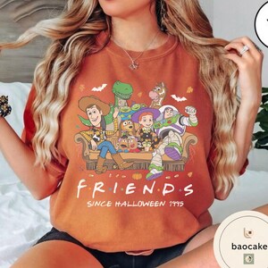 May include: A rust-coloured crewneck sweatshirt featuring a graphic print of Toy Story characters sitting on a sofa. The text below reads "F R I E N D S" and "SINCE HALLOWEEN 1995". The design includes small pumpkins, bats, and spiderwebs.