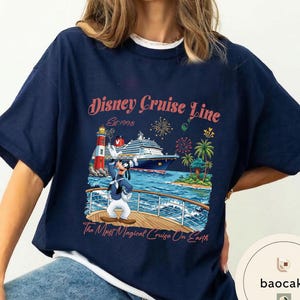 K&ouml;nnte beinhalten: Marineblaues T-Shirt mit einem Disney Cruise Line-Motiv. Das Design zeigt ein Kreuzfahrtschiff, einen Leuchtturm, Palmen, Feuerwerk und Goofy. Der Text auf dem Shirt lautet "Disney Cruise Line Est. 1998" und "The Most Magical Cruise On Earth."