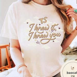K&ouml;nnte beinhalten: Ein cremefarbenes Kurzarm-T-Shirt mit dem Text "TS I knew it, I knew you" in einer braunen Schriftart im Seil-Stil. Das Design enth&auml;lt kleine braune Herzen, hellbeige Wolkenformen und eine kleine florale Grafik mittig auf der Brust.