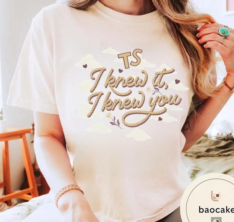 K&ouml;nnte beinhalten: Ein cremefarbenes Kurzarm-T-Shirt mit dem Text "TS I knew it, I knew you" in einer braunen Schriftart im Seil-Stil. Das Design enth&auml;lt kleine braune Herzen, hellbeige Wolkenformen und eine kleine florale Grafik mittig auf der Brust.