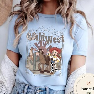 Pu&ograve; includere: T-shirt azzurra con un design del West selvaggio. Il design include una cowgirl dei cartoni animati, un cavallo e la scritta "Wild West". Il design include anche un ferro di cavallo e un fiocco. La maglietta ha maniche corte e girocollo.