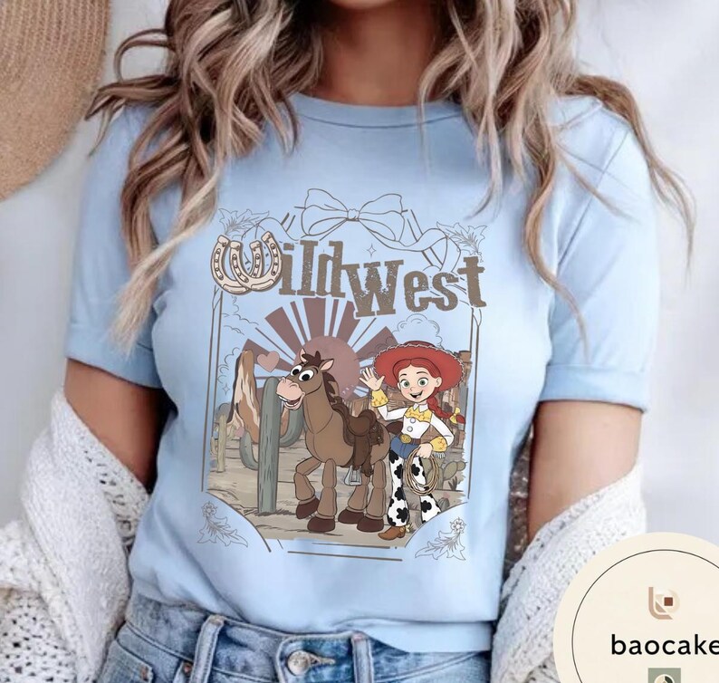 Pu&ograve; includere: T-shirt azzurra con un design del West selvaggio. Il design include una cowgirl dei cartoni animati, un cavallo e la scritta "Wild West". Il design include anche un ferro di cavallo e un fiocco. La maglietta ha maniche corte e girocollo.