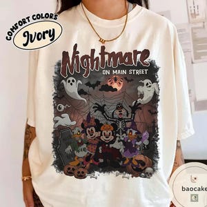 May include: Ivory-colored Comfort Colors t-shirt featuring a Halloween-themed graphic. The graphic includes the text "Nightmare on Main Street" with cartoon characters, ghosts, bats, and a full moon. The design has a spooky, festive vibe.