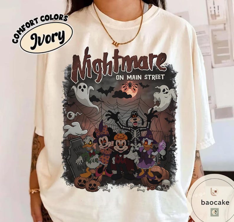 May include: Ivory-colored Comfort Colors t-shirt featuring a Halloween-themed graphic. The graphic includes the text "Nightmare on Main Street" with cartoon characters, ghosts, bats, and a full moon. The design has a spooky, festive vibe.