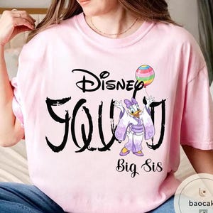 Puede incluir: Camiseta rosa claro con la palabra "Disney" encima de "Young" en negro. Un dibujo animado de Daisy Duck con un kimono sosteniendo un globo est&aacute; en la camiseta. Las palabras "Big Sis" est&aacute;n debajo.