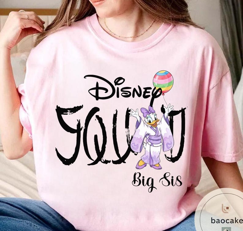Puede incluir: Camiseta rosa claro con la palabra "Disney" encima de "Young" en negro. Un dibujo animado de Daisy Duck con un kimono sosteniendo un globo est&aacute; en la camiseta. Las palabras "Big Sis" est&aacute;n debajo.