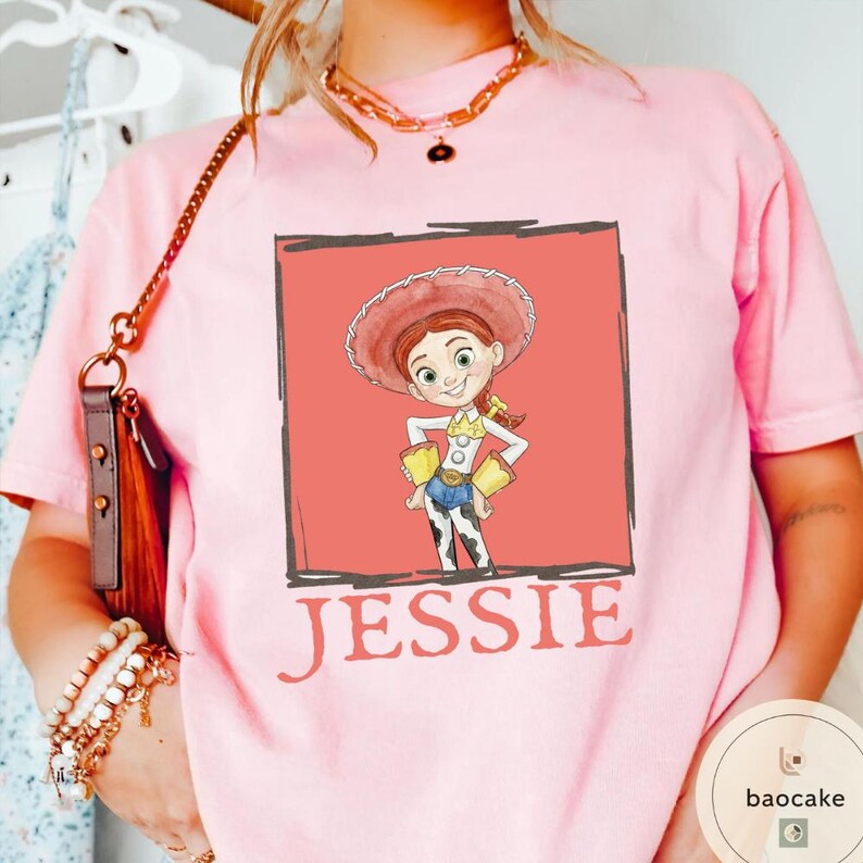 K&ouml;nnte beinhalten: Ein hellrosa T-Shirt mit einer Grafik der Figur Jessie aus Toy Story in einem korallenfarbenen quadratischen Rahmen. Die Figur tr&auml;gt einen roten Cowgirl-Hut, ein gelbes Hemd mit wei&szlig;en Manschetten, eine blaue Hose und Chaps mit Kuhfleckenmuster. Der Name JESSIE ist darunter aufgedruckt.