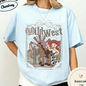 Pu&ograve; includere: T-shirt azzurra con grafica Wild West. La grafica include una cowgirl dei cartoni animati, un cavallo e la scritta "Wild West". Il design incorpora un ferro di cavallo ed elementi del paesaggio desertico. La maglietta ha scollo tondo e maniche corte.