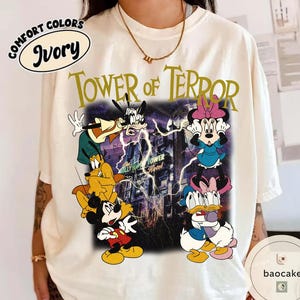 May include: Ivory-colored t-shirt featuring a graphic of Disney characters, including Mickey Mouse, Goofy, and Minnie Mouse, in front of the Hollywood Tower Hotel. The text "TOWER OF TERROR" is printed above the graphic.