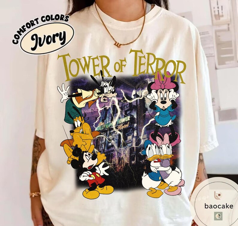 May include: Ivory-colored t-shirt featuring a graphic of Disney characters, including Mickey Mouse, Goofy, and Minnie Mouse, in front of the Hollywood Tower Hotel. The text "TOWER OF TERROR" is printed above the graphic.