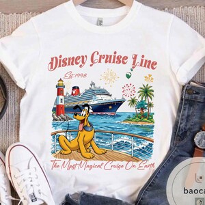 K&ouml;nnte beinhalten: Wei&szlig;es T-Shirt mit einer farbenfrohen Grafik eines Kreuzfahrtschiffs, eines Leuchtturms und Pluto. Der Text "Disney Cruise Line Est.1998" ist in einer dekorativen Schriftart dargestellt. Der Satz "The Most Magical Cruise On Earth" ist ebenfalls enthalten.