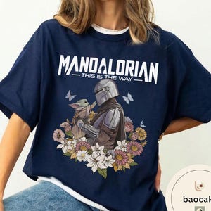 Puede incluir: Camiseta azul marino con el texto blanco "MANDALORIAN - THIS IS THE WAY". El dise&ntilde;o muestra al Mandaloriano y a Grogu, rodeados de flores y mariposas.