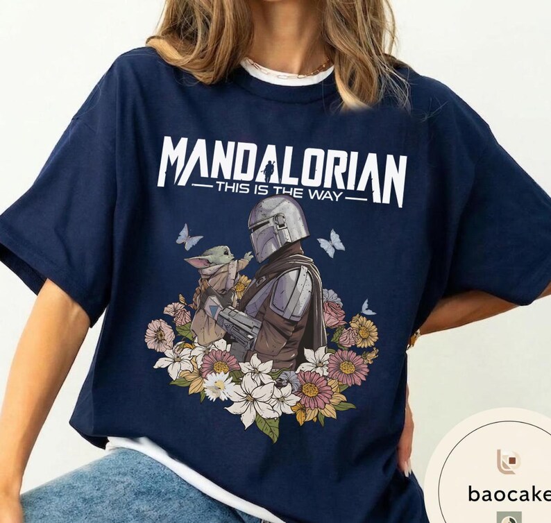 Puede incluir: Camiseta azul marino con el texto blanco "MANDALORIAN - THIS IS THE WAY". El dise&ntilde;o muestra al Mandaloriano y a Grogu, rodeados de flores y mariposas.