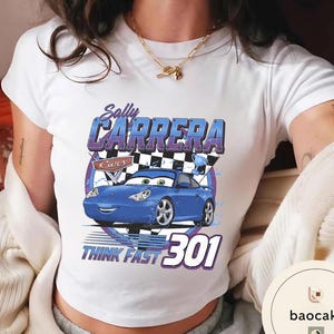 Puede incluir: Camiseta blanca con un dise&ntilde;o de coche de dibujos animados azul, con el texto "Sally Carrera" y "Think Fast 301". El dise&ntilde;o incluye una bandera a cuadros y un trofeo. La camiseta es de estilo corto.