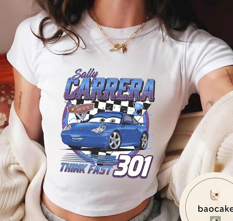 Puede incluir: Camiseta blanca con un dise&ntilde;o de coche de dibujos animados azul, con el texto "Sally Carrera" y "Think Fast 301". El dise&ntilde;o incluye una bandera a cuadros y un trofeo. La camiseta es de estilo corto.