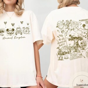 May include: Cream-colored t-shirt with a green graphic design. The front features "Animal Kingdom" text and themed illustrations. The back displays a detailed map with the text "Adventure Awaits!" and "Peak Challenge."