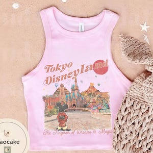May include: A light pink tank top with the words "Tokyo Disneyland" in a vintage font. The shirt features a colorful graphic of a Disney park scene with Mickey Mouse. The text "The Kingdom of Dreams & Magic" is below the graphic.