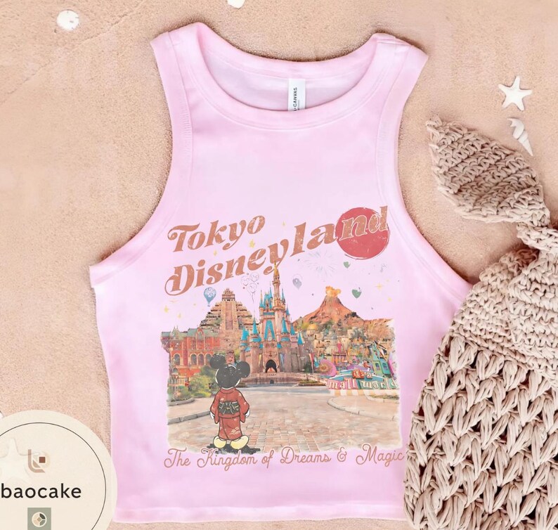 May include: A light pink tank top with the words "Tokyo Disneyland" in a vintage font. The shirt features a colorful graphic of a Disney park scene with Mickey Mouse. The text "The Kingdom of Dreams & Magic" is below the graphic.