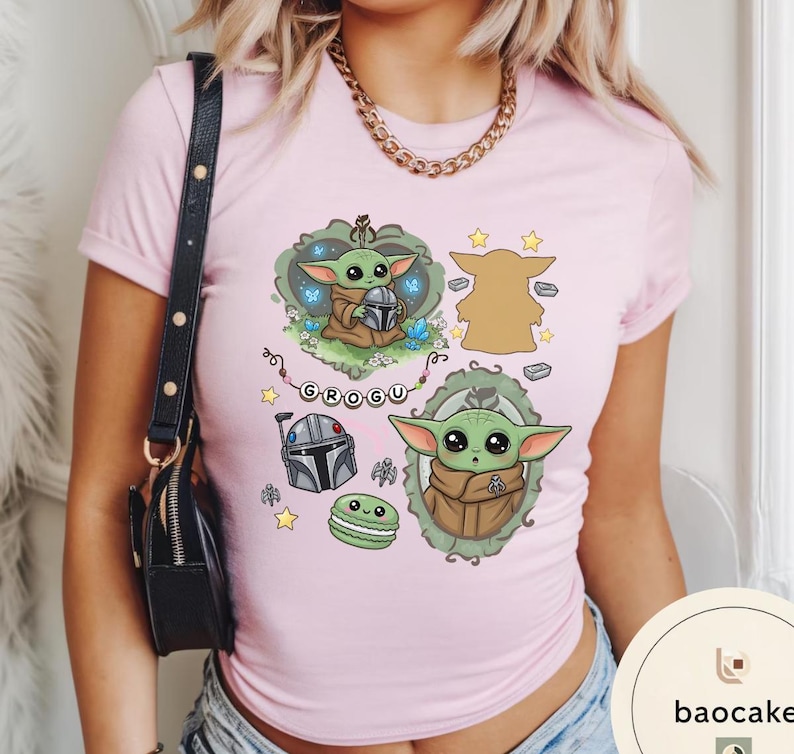 May include: A light pink t-shirt featuring a graphic print of the character Grogu in various poses, a Mandalorian helmet, a green macaron, and a string of beads spelling GROGU. The design includes decorative frames, small stars, and blue butterflies.