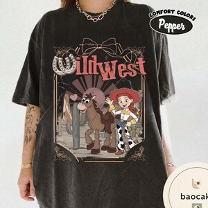 Pu&ograve; includere: T-shirt grigio scuro con una grafica del West selvaggio con una cowgirl dei cartoni animati, un cavallo e la scritta "Wild West". Il design include un ferro di cavallo e un paesaggio desertico. La maglietta ha un'etichetta Comfort Colors Pepper.