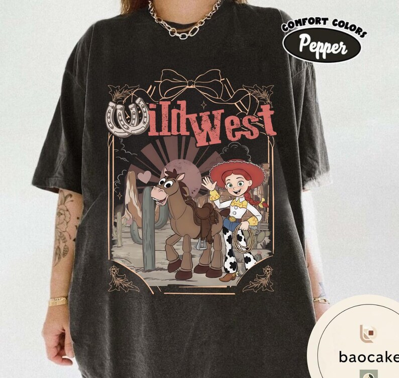 Pu&ograve; includere: T-shirt grigio scuro con una grafica del West selvaggio con una cowgirl dei cartoni animati, un cavallo e la scritta "Wild West". Il design include un ferro di cavallo e un paesaggio desertico. La maglietta ha un'etichetta Comfort Colors Pepper.