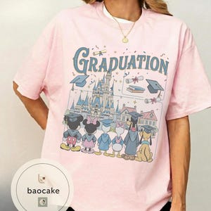 May include: A light pink t-shirt featuring a graduation design with the word "Graduation" in blue script. The graphic includes cartoon characters, graduation caps, a castle, and scrolls. The shirt has short sleeves.
