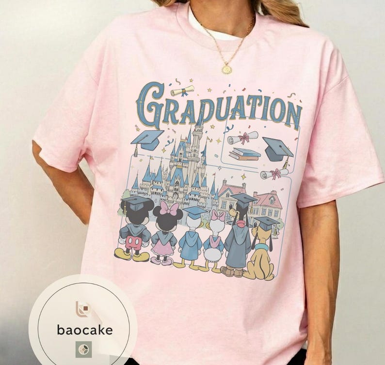May include: A light pink t-shirt featuring a graduation design with the word "Graduation" in blue script. The graphic includes cartoon characters, graduation caps, a castle, and scrolls. The shirt has short sleeves.