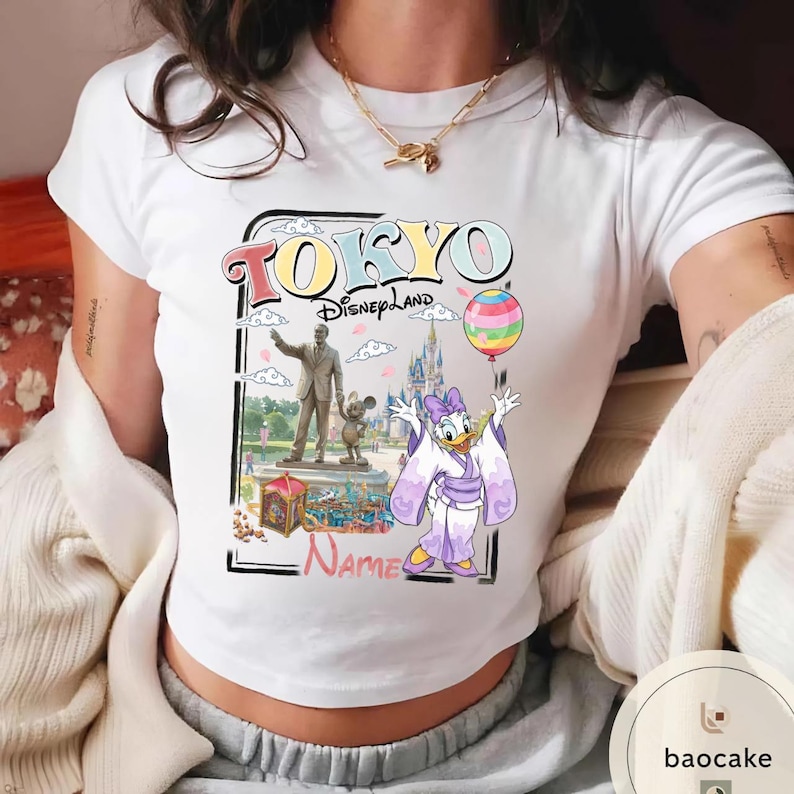 Puede incluir: Camiseta blanca con un gr&aacute;fico colorido de Tokyo Disneyland. El dise&ntilde;o incluye el texto "TOKYO Disneyland" con una imagen de Daisy Duck con un kimono, una estatua y un castillo. Tambi&eacute;n se incluye un globo de arco&iacute;ris.