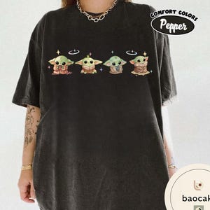 May include: Black t-shirt featuring four cartoon characters with large eyes and pointed ears. Each character is wearing a different outfit. The text "Comfort Colors Pepper" is in a white oval on the upper right.