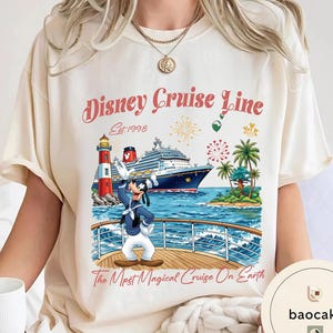 K&ouml;nnte beinhalten: Cremefarbenes T-Shirt mit einem Disney Cruise Line-Motiv. Das Design zeigt ein Schiff, Goofy, einen Leuchtturm und Palmen. Der Text lautet "Disney Cruise Line Est. 1998" und "The Most Magical Cruise On Earth."