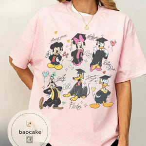 May include: A light pink t-shirt featuring a graphic of Disney characters Mickey Mouse, Minnie Mouse, Donald Duck, Daisy Duck, Goofy, and Pluto in graduation attire. The shirt has the text "Mickey Mouse" and character signatures.