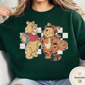 May include: Dark green sweatshirt featuring two cartoon characters, one in a tiger costume holding a pumpkin, and the other with a backpack. The characters are set against a white and black checkered background.