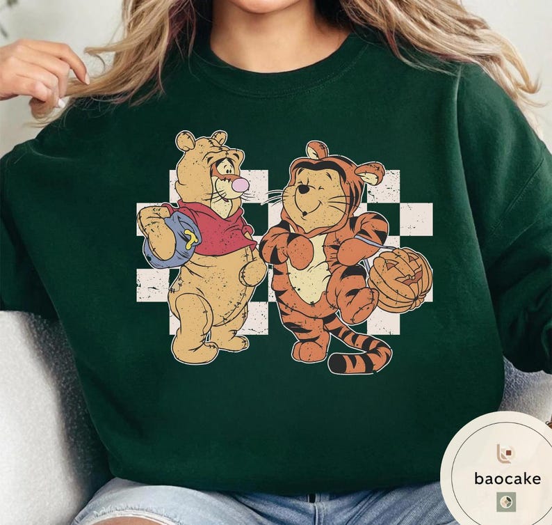 May include: Dark green sweatshirt featuring two cartoon characters, one in a tiger costume holding a pumpkin, and the other with a backpack. The characters are set against a white and black checkered background.