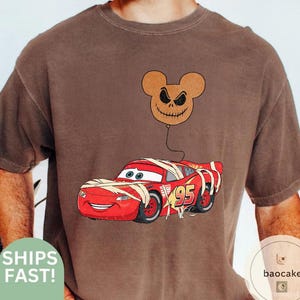 May include: Brown t-shirt featuring a red cartoon race car wrapped in bandages with the number 95. A brown balloon with a Jack Skellington face is attached to the car. The shirt has a vintage look.