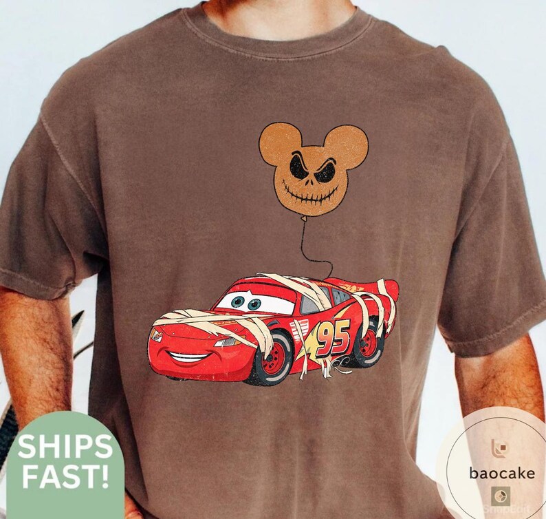 May include: Brown t-shirt featuring a red cartoon race car wrapped in bandages with the number 95. A brown balloon with a Jack Skellington face is attached to the car. The shirt has a vintage look.