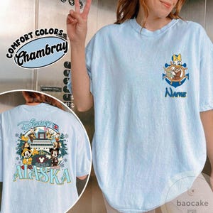 May include: Light blue t-shirt with a Disney-themed graphic. The front features a nautical design with a character and the word "Name". The back displays a Disney Alaska 2024 design with Mickey and Minnie Mouse. The shirt is labeled "Comfort Colors Chambray".