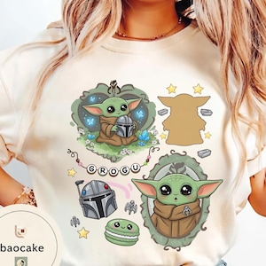 May include: A cream-colored t-shirt featuring a graphic print of Grogu, a Mandalorian helmet, a green macaron, and stars. The design includes the text "GROGU" on a string of beads, surrounded by decorative frames and small icons in shades of green, brown, and grey.