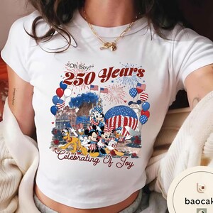 Puede incluir: Camiseta blanca con un gr&aacute;fico colorido que celebra 250 a&ntilde;os. El dise&ntilde;o incluye a Mickey Mouse, Minnie Mouse, Donald Duck y otros personajes de Disney, junto con fuegos artificiales, globos y banderas americanas. El texto incluye "Oh Boy!" y "Celebrating of Joy."