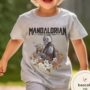 Puede incluir: Camiseta gris claro con el texto "MANDALORIAN - THIS IS THE WAY -" sobre un gr&aacute;fico del Mandaloriano y Grogu rodeados de flores y mariposas. La camiseta es de manga corta y cuello redondo.