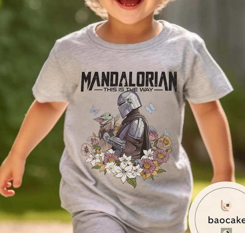 Puede incluir: Camiseta gris claro con el texto "MANDALORIAN - THIS IS THE WAY -" sobre un gr&aacute;fico del Mandaloriano y Grogu rodeados de flores y mariposas. La camiseta es de manga corta y cuello redondo.